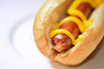Closeup of a cooked hot dog in a plain soft bun with mustard on a white plate.