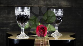 Red Rose and Wine Glasses Resting On Acoustic Guitar With Wooden Background