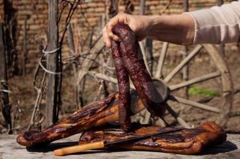 Smoked Dried Sausages Held By Woman's Hand With Smoked Bacon On A Rustic Wooden Surface. Delicious Domestic Food