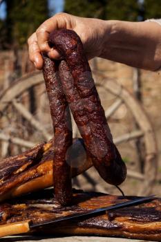 Smoked Dried Sausages Held By Woman's Hand With Smoked Bacon On A Rustic Wooden Surface. Delicious Domestic Food