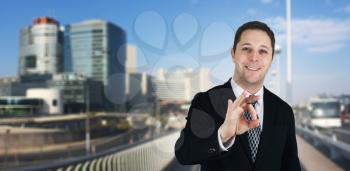Businessman Smiling and Feeling Happy Giving Ok Sign With Business City and Corporate Buildings In Background