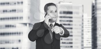 Businessman Talking On The Phone And Pointing Index Finger Towards Camera With Business City and Corporate Buildings In Background