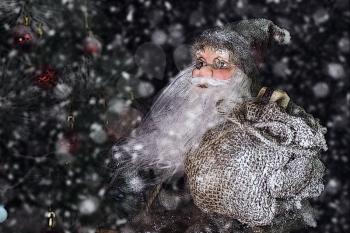 Santa Claus Outdoors Beside Christmas Tree in Snowfall Carrying Gifts to Children. Merry Christmas & New Year's Eve concept