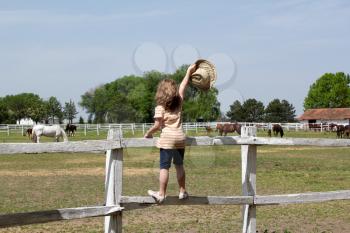 little girl with hat in hand standing on a corral fence