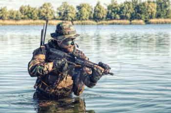 Bearded soldier of special forces in action during river raid in the jungle terrain. He is waist deep in the water and mud and ready to meet enemy, survive and fight in agressive hostile environment