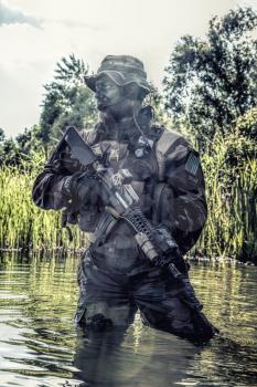 Bearded soldier of special forces in action during river raid in the jungle terrain. He is waist deep in the water and mud and ready to meet enemy, survive and fight in agressive hostile environment
