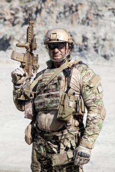 Half length location shot of big muscular soldier in field uniforms with rifle in the desert among rocks