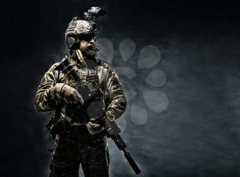 Bearded soldier in Combat Uniforms with weapon, plate carrier and combat helmet are on. Studio shot, dark background