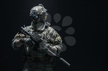 Army soldier in Combat Uniforms with assault rifle, plate carrier and combat helmet are on, Shemagh Kufiya scarf on his neck. Studio shot, dark background