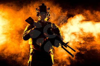 Army soldier in Combat Uniforms with machine gun, plate carrier and combat helmet are on. Studio contour silhouette shot, backlight, dark glowing smoke background