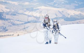 Army servicemen in winter camo somewhere in the mountains. Walking moving across snow desert despite bad weather wind and cold, holding weapons - assault and sniper rifle. Full body portrait