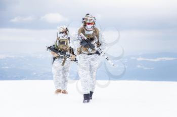 Army servicemen in winter camo somewhere in the mountains. Walking moving across snow desert despite bad weather wind and cold, holding weapons - assault and sniper rifle. Full body portrait
