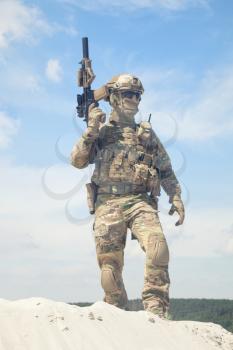 Man in military camouflage uniform and mask, equipped tactical ammunition, standing on sand dune with service rifle replica in hands, cloudy sky on background. Airsoft player taking part in war games