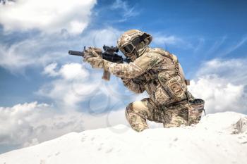Man in military camouflage uniform with service rifle replica, standing on top of sand dune with cloudy sky on background, imitating U.S. army special forces shooter during airsoft war games in desert