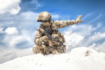 Army soldier or airsoft player in camo uniform and helmet, armed assault rifle, standing on knee on sand dune, looking back and showing halt or stop hand signal. Nonverbal communication on battlefield