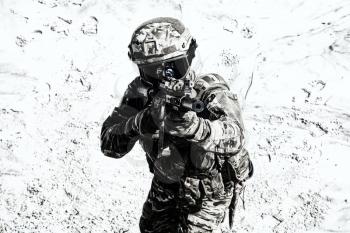 Airsoft player in camo combat uniform, protected with helmet and ballistic glasses, aiming with optical sight on army service rifle replica in sandy area desaturated shoot. War games participant