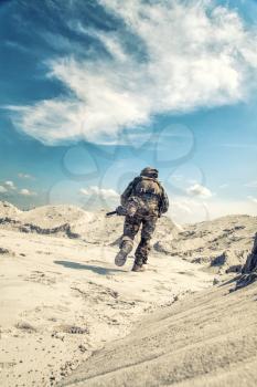 Man in military camouflage uniform with airsoft weapon bending down and running during armed conflict, military battle imitation in sandy or desert area. Airsoft player under fire searching cover