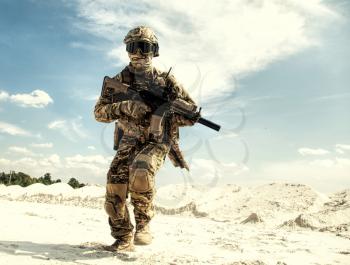 Airsoft player in camouflage uniform protected with helmet and ballistic goggles running with army service rifle replica in sandy area during military operation, war collisions or battle simulation