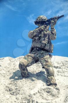 Airsoft military games participant in U.S. army soldier camouflage uniform, helmet, tactical goggles and load carrier, aiming with optical sight on carbine replica while sliding down from sand dune