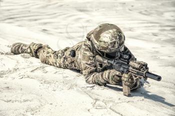Man wearing camo uniform, equipped with military ammunition, lying on sand and shooting with service rifle replica while playing war game, taking part in desert battle simulation during airsoft event
