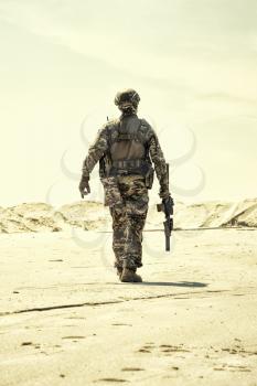 Military contractor, army infantry or rifleman in camouflage uniform and helmet patrolling territory in desert. Airsoft player with real firearm replica walking in sandy area desaturated, back view
