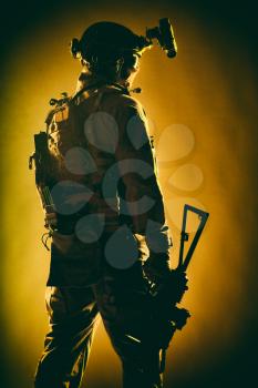 Army special forces soldier in tactical ammunition, night vision device on helmet, standing with lowered service rifle in hand low key, back view, studio shoot with red backlight on fabric background