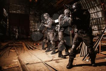 Police special operations anti-narcotics or counter-terrorism team, quick reaction tactical group in black uniforms and masks, armed with semi-automatic rifles moving with caution in abandoned hangar