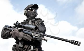 Closeup portrait of police special operations sniper in black blank uniforms and mask, body armor, ballistic goggles and headphones standing on sky background, holding sniper rifle with optical sight