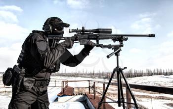 Sniper of police special operations tactical group in black uniforms, ballistic glasses and headphones, aiming with telescopic optical sight on sniper rifle mounted on tripod, over shoulder back view