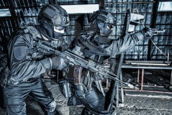 Police special forces, counter-terrorism tactical team fighters, private security company guards aiming guns while moving forward under cover of ballistic shield on anti narcotics raid, desaturated