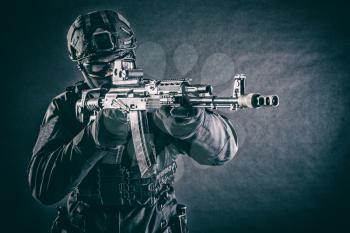 Police tactical team member, army special forces, private security company fighter in black uniform, helmet and mask aiming with collimator sight on assault rifle, toned in blue, low key studio shoot