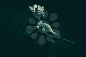Half length, low key studio shoot of army soldier, marine infantryman in mask, camo uniform, equipped modern ammunition, armed service rifle standing in darkness with night vision device on helmet