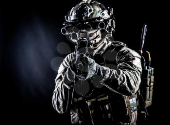 Soldier of special forces in camo combat uniform, load carrier, helmet, equipped night sight goggles, tactical radio headset, aiming assault rifle with collimator sight in camera, low key studio shoot