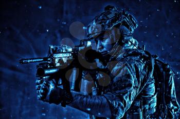 Anti terrorist squad fighter, army special forces rifleman in battle uniform, tactical radio headset, backpack, body armor, aiming service rifle with collimator sight, sneaking in darkness under rain