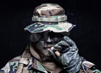 Brutal and serious commando soldier, army special forces veteran, in camouflage battle uniform, boonie hat, black paint on bearded face, combat knife in shoulder holder, smoking cigar, studio portrait
