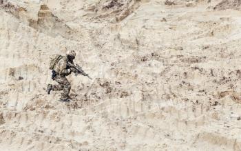Army soldier or commando with assault rifle in ocp camouflage combat uniform, wearing helmet and with backpack on back running fast in sands. Special military operation in dessert, war on Middle East