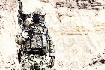 Army soldier, private military company fighter with hidden identity in battle uniform, helmet, body armor and radio headset, standing with service rifle in rocky area
