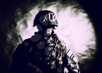 Low key studio portrait of private security service contractor, army infantry rifleman, US marine raider in helmet, sunglasses, camouflage uniform posing with weapon on black background with backlight