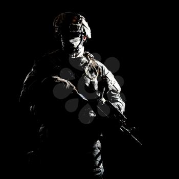 US marine raider in combat uniform with hidden face, armed with assault carbine low key, high contract studio shot on black background. Equipped army soldier standing in darkness with weapon in hands