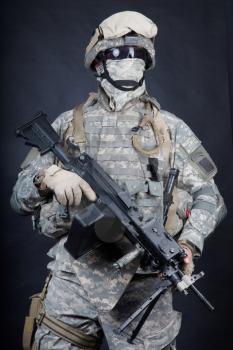 Marine with hiding identity, protected with helmet and body armour in digital camouflage combat uniform posing with light machine gun equipped infrared laser aiming light isolated on black background
