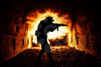 Silhouette of post apocalyptic soldier or survivor shooting with automatic firearm weapon in dark dungeon, city sewage tunnels, or underground bunker with explosion fire and smoke on background
