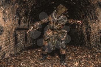 Post apocalyptic mutant creature or survivor in tatters and gas mask jumps out of darkness and attacking with handmade machete in abandoned tunnel, frightening dungeon or city old sewage collector