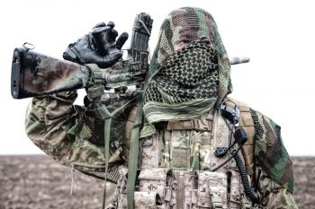 Army marksman, airsoft player in camouflage uniform and load carrier, masking cape on head, armed service rifle with optical sight, hiding face with shemagh, standing on field, looking into distance