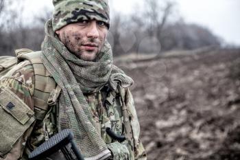 Portrait of soldier, modern combatant with dirty face, firearm replica, wearing camouflage uniform, beanie and masking cape on neck, standing on field, carrying backpack on march