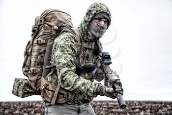 Irregular military mercenary, commando saboteur in camo jacket, wearing hood on head, armed service rifle, carrying tactical backpack during march, looking on distance