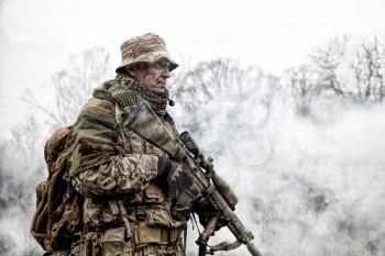 Elite commando fighter, private military company mercenary, army special forces veteran in camouflage uniform, shemagh and bonnie, equipped radio headset standing with sniper rifle in clouds of smoke