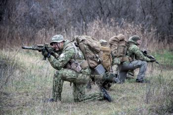 Airsoft, strikeball players team, commando group moving cautiously in forest area, kneeling and looking around, covering comrades, controlling sectors. Commander showing halt or stop hand signal