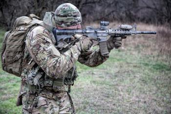 Army infantry, special forces fighter, commando shooter in camouflage uniform, beanie, carrying backpack, aiming service rifle, shooting on enemy in autumn forest, observing area with collimator sight