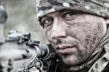 Close-up portrait of army infantryman, modern warfare combatant, young soldier with dirty, unshaven face, in camo bennie hat, aiming service rifle, controlling area with gunfire, looking in camera
