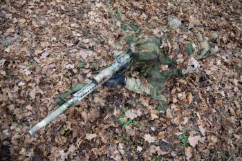 Army elite forces sniper, tactical group marksman, airsoft player lying on ground in forest, hiding in autumn foliage, covering himself with camouflage cape, searching targets through optical sight